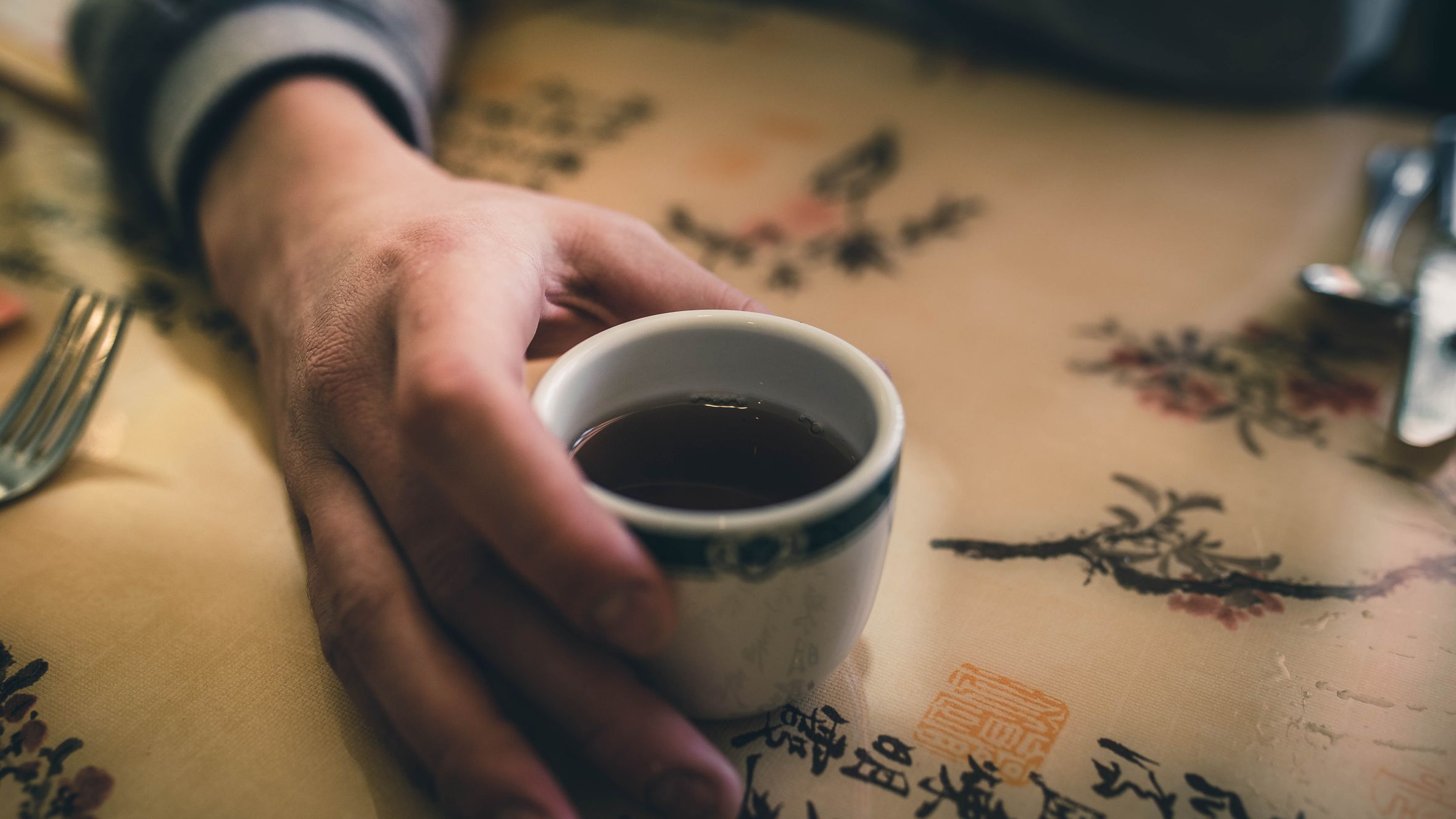A close-up of a hand holding a cup of hot tea on a table with artistic Asian prints.