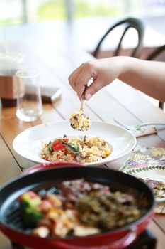 A close-up of a hand lifting a spoonful of colorful fried rice with diverse ingredients from a plate.