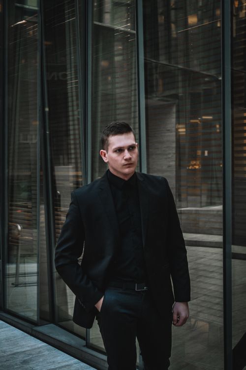Free Young man in sleek dark suit stands confidently by a modern glass building facade. Stock Photo
