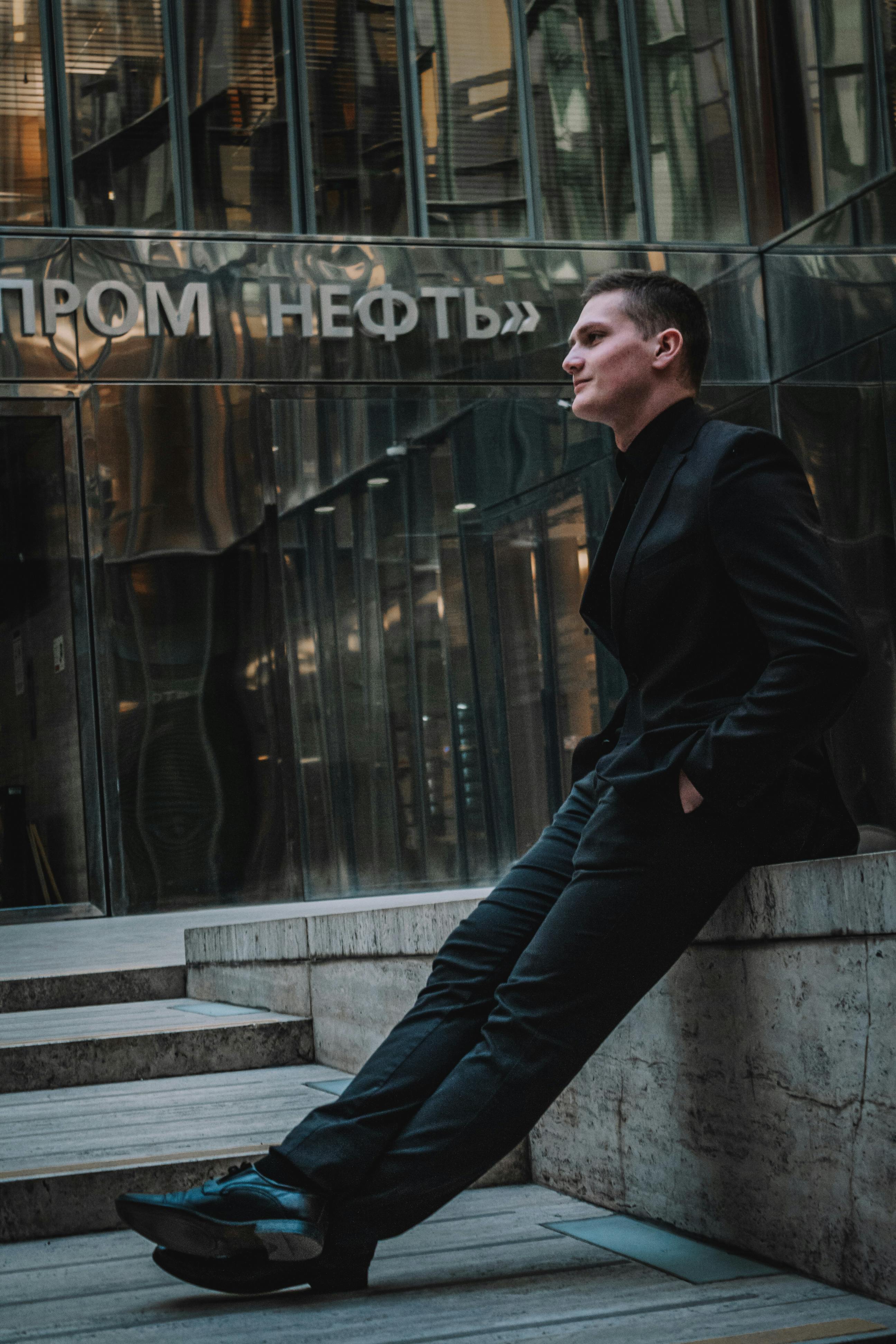 A young man in a black suit leans casually against a sleek building, exuding modern elegance.