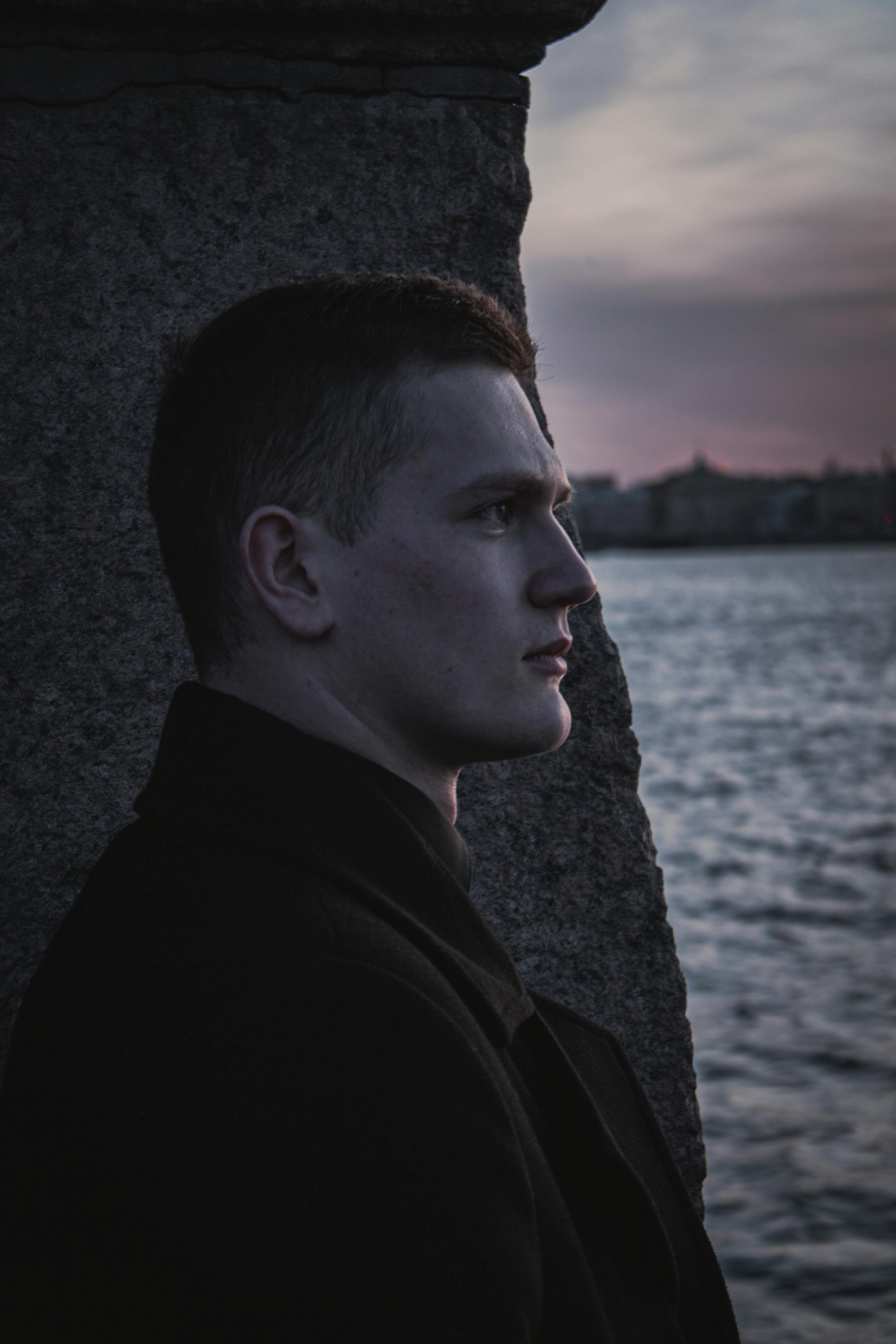 A contemplative man in a black coat gazes over the sea at dusk, evoking a serene mood.