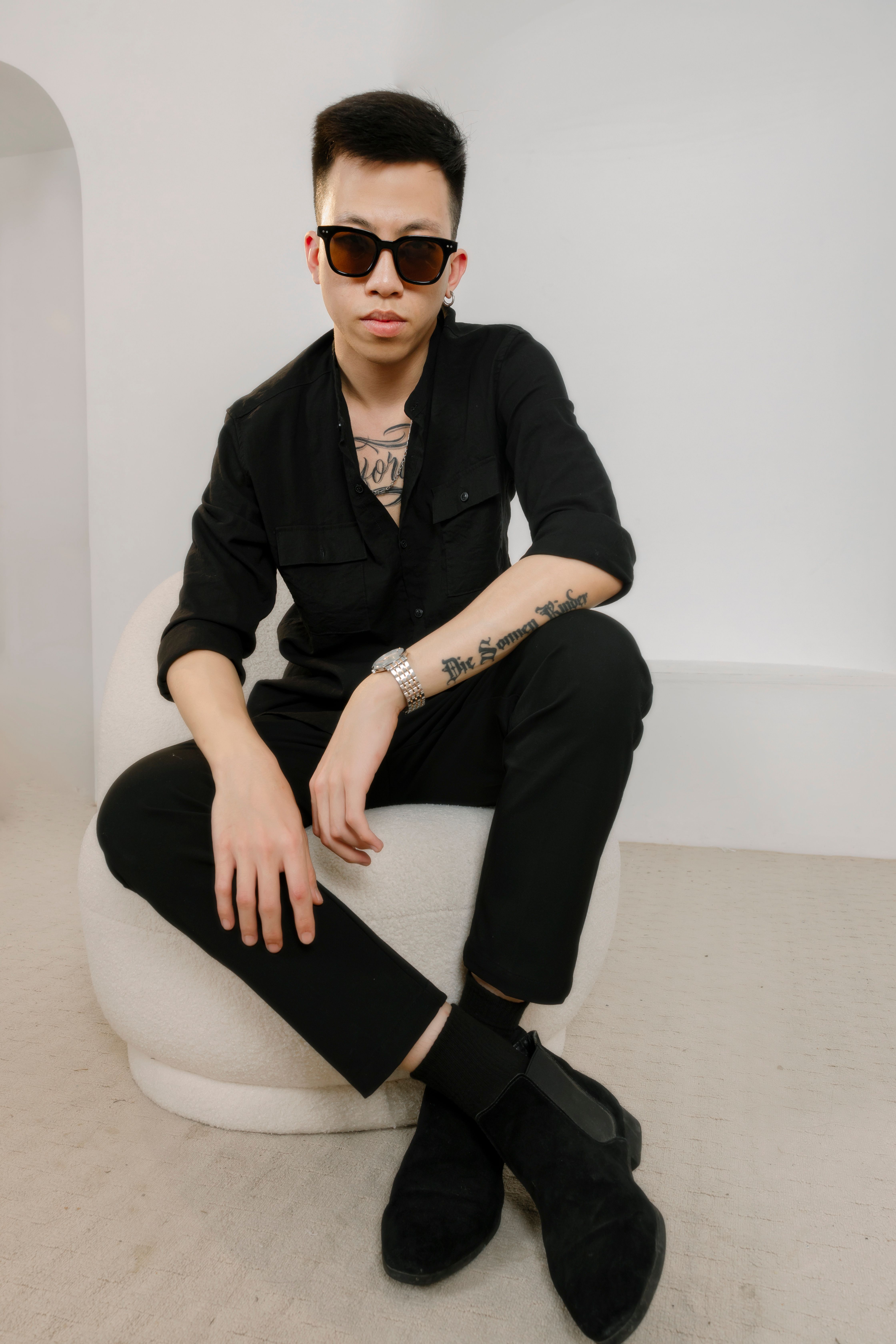 Portrait of a fashionable man with tattoos, wearing sunglasses and black attire, sitting indoors.
