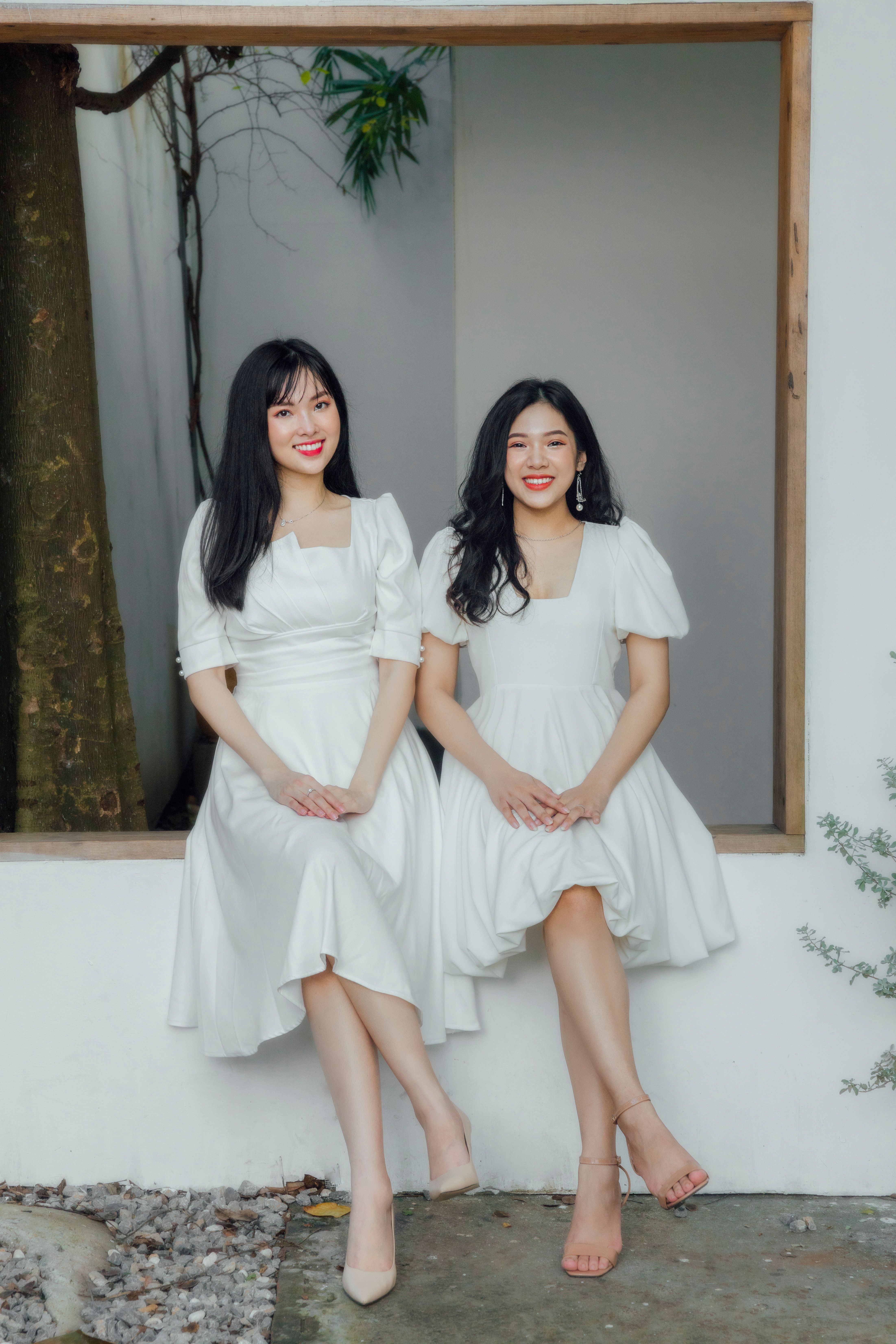 Two women sitting on a windowsill wearing white dresses, showing fashion and friendship.