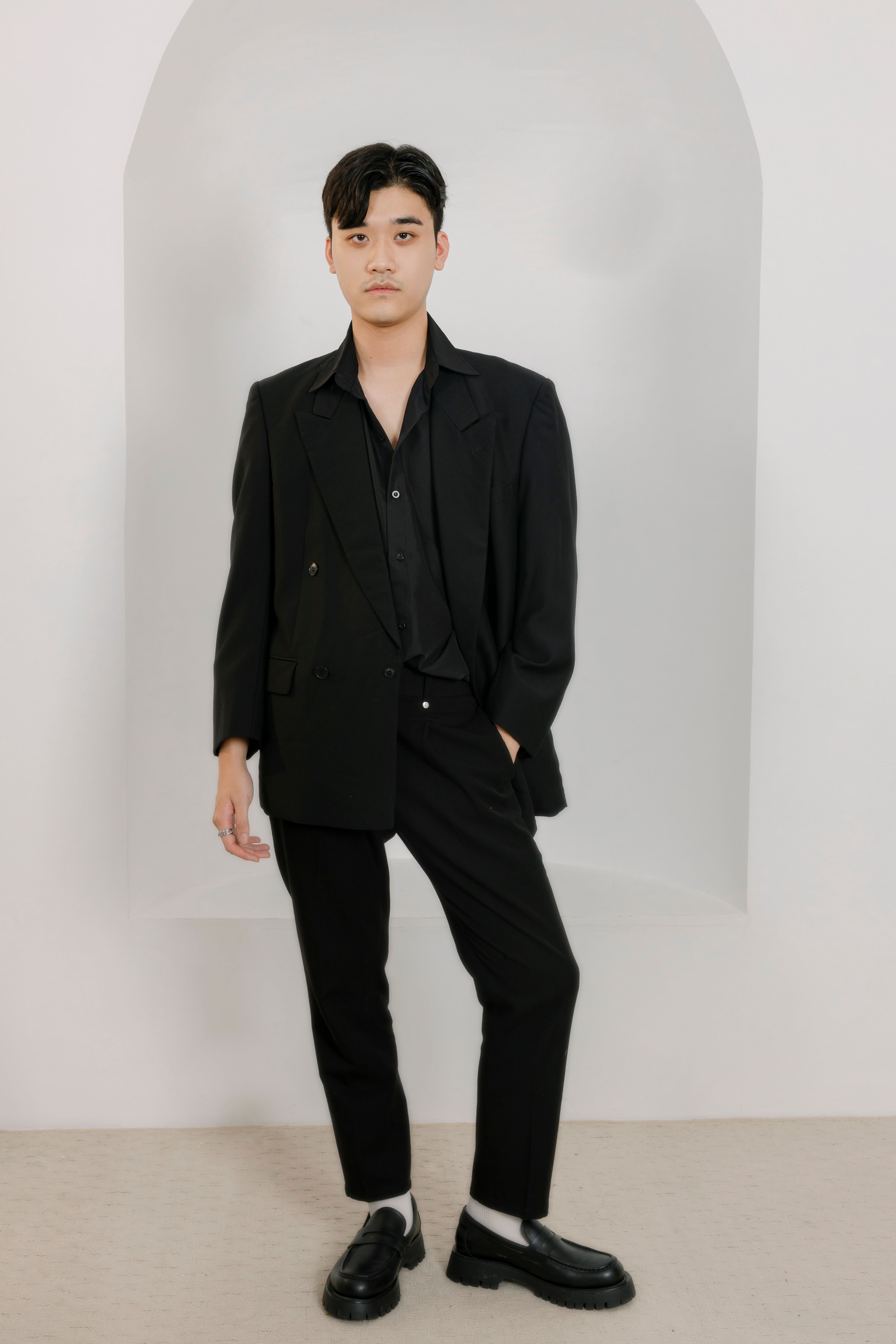 Portrait of a stylish Asian man in black suit standing confidently in a studio setting.