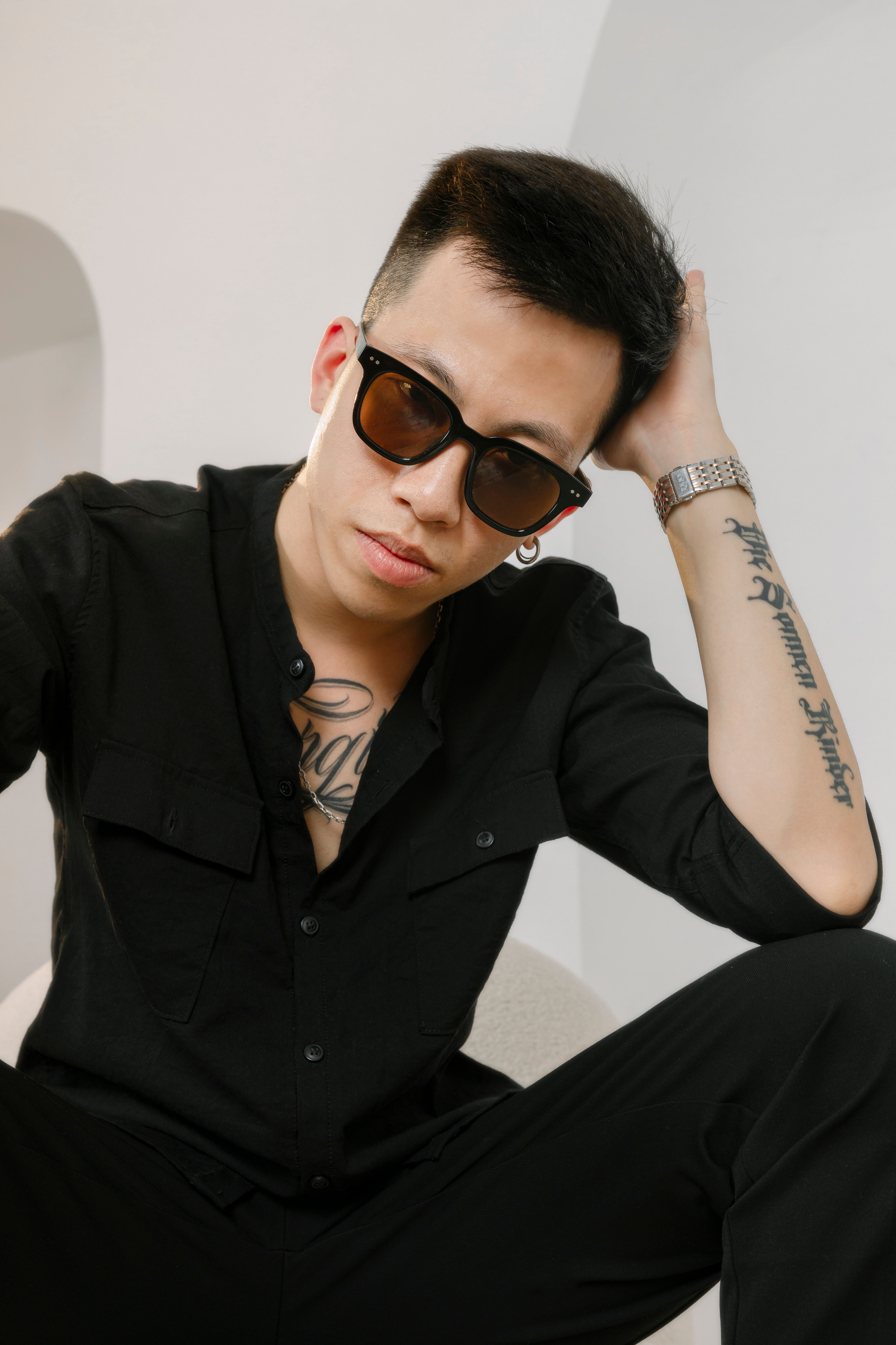Trendy young man in sunglasses and black clothes posing casually indoors.