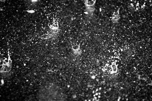 Monochrome image of raindrops splashing on a textured concrete surface, capturing dynamic water movement.