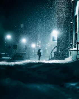 A person stands on a snow-covered street at night during heavy snowfall, surrounded by dim lights.