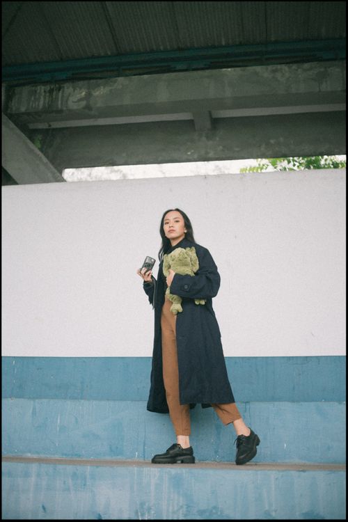 Free Young woman in trench coat with a camera and stuffed toy, standing before a white and blue wall. Stock Photo