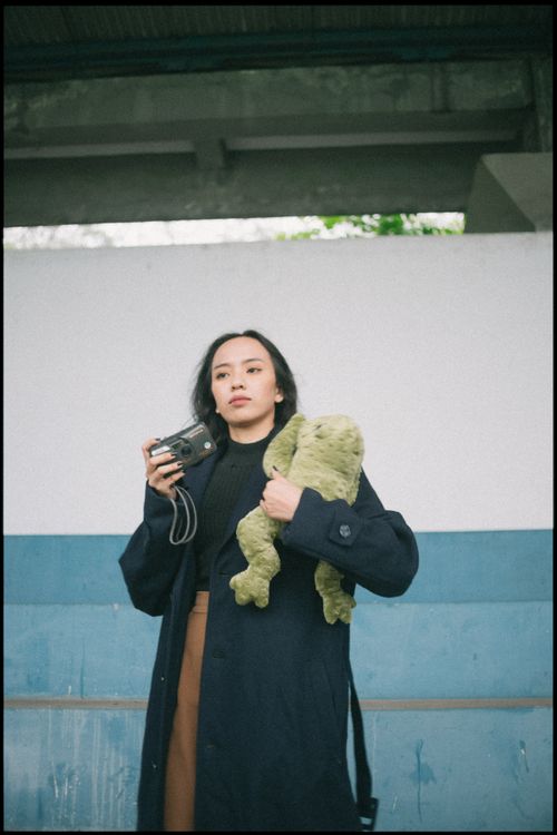 Free Stylish young woman in a trench coat holding a camera and stuffed toy, standing against a concrete wall. Stock Photo