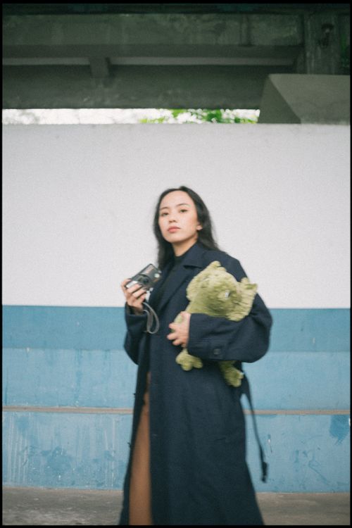 Free A woman in a long coat holding a camera and teddy bear, posing outdoors confidently. Stock Photo