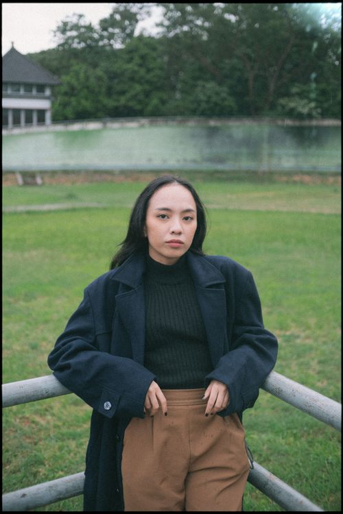 Free A fashionable young woman stands confidently by a railing outdoors, wearing a trench coat and brown pants. Stock Photo