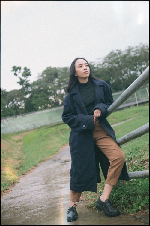 Free Asian woman in a black coat leaning on railing in a green park area. Stock Photo