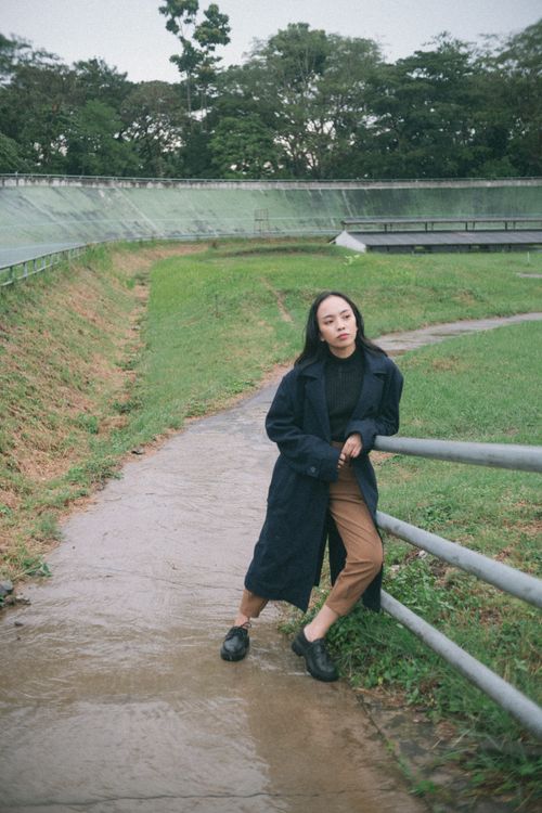 Free Young woman in trench coat leans on railing by a wet pathway outdoors. Stock Photo