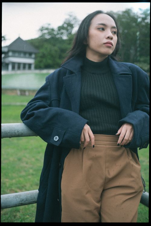 Free Stylish young woman in a dark coat leaning on a fence in a park setting, exuding a modern urban vibe. Stock Photo