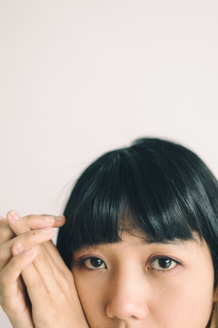 Close-up image of a woman with dark hair and bangs, hands together against a soft background.