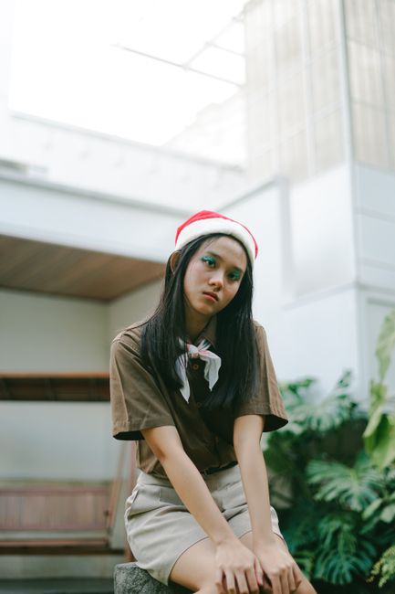 A young woman wearing a Christmas hat and brown shirt sits indoors, looking serene and thoughtful.