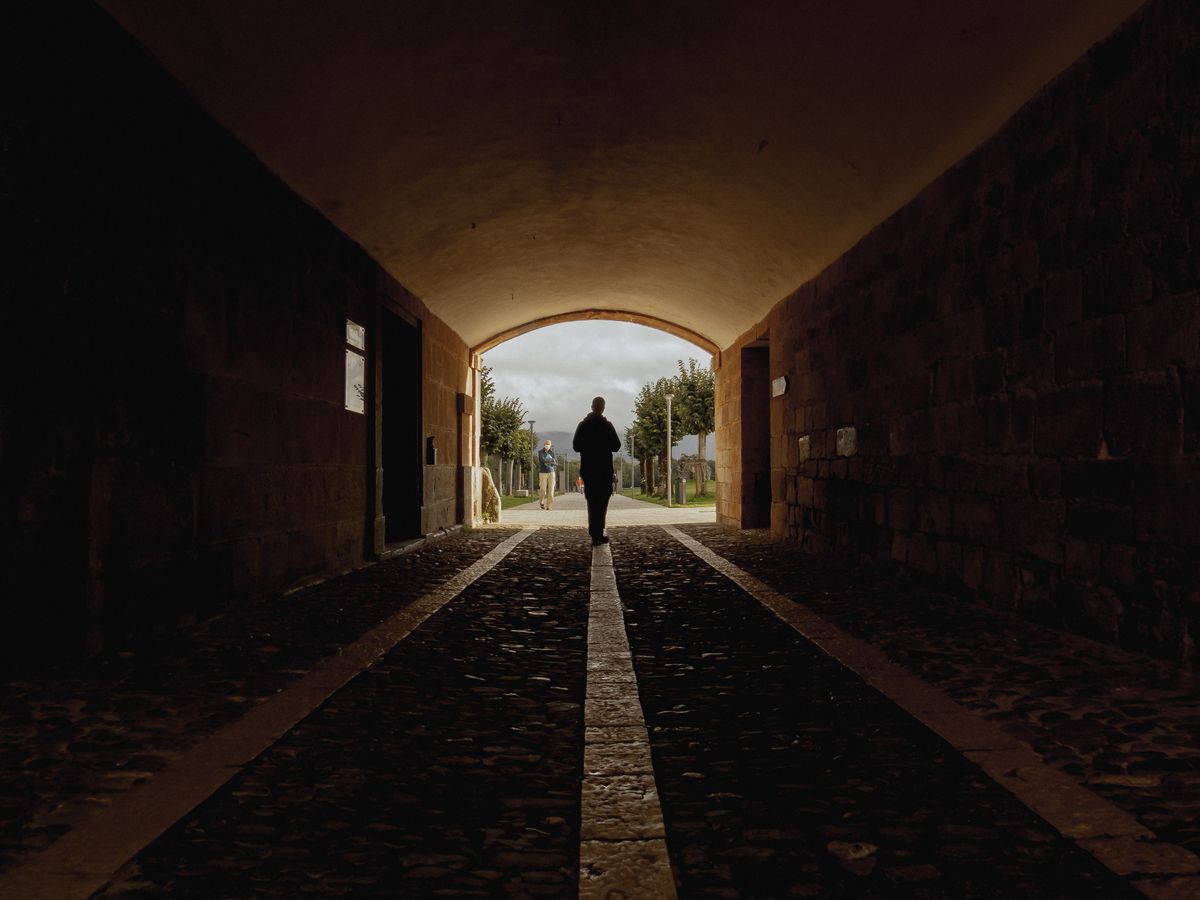 A lone figure walking a dark stone passage toward a lit arch — photo by Berta Raspall on Pexels