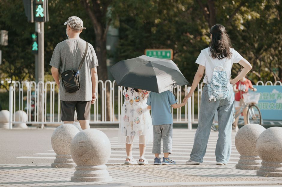 A family waits at a crosswalk with children holding an umbrella on a sunny day.