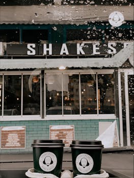 Rainy day scene of two takeaway coffee cups in front of a vintage milkshake shop.
