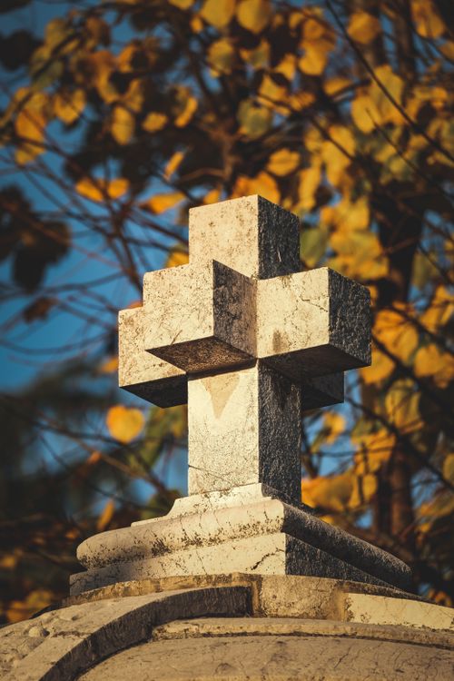 Free Close-up of a stone cross atop a tomb with autumn leaves in the background under clear blue sky. Stock Photo