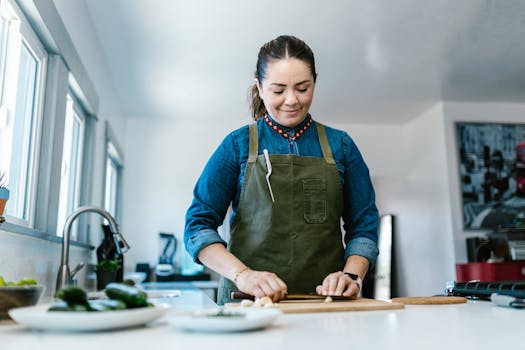 Woman in a kitchen chopping vegetables on a cutting board, wearing a green apron with focus on the preparation.