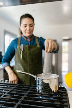 Woman seasoning food in a modern kitchen, wearing an apron by a gas stove.