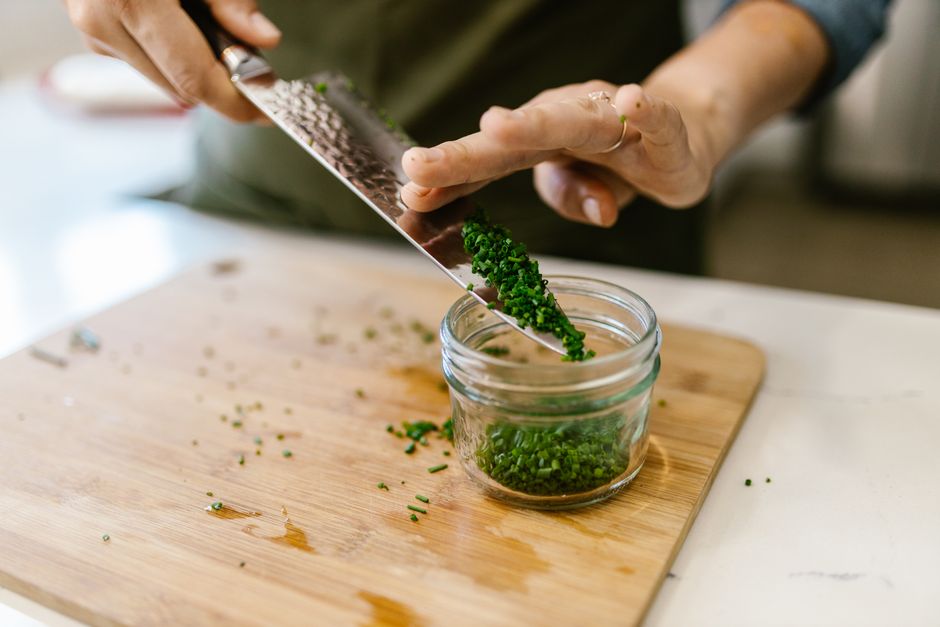 A person chops fresh chives into a jar on a wooden cutting board, showcasing food preparation.