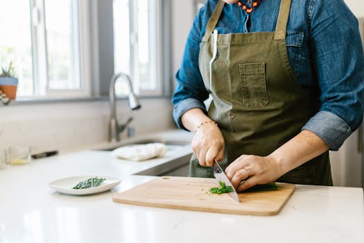 Woman in apron chops herbs on wooden board in bright kitchen.