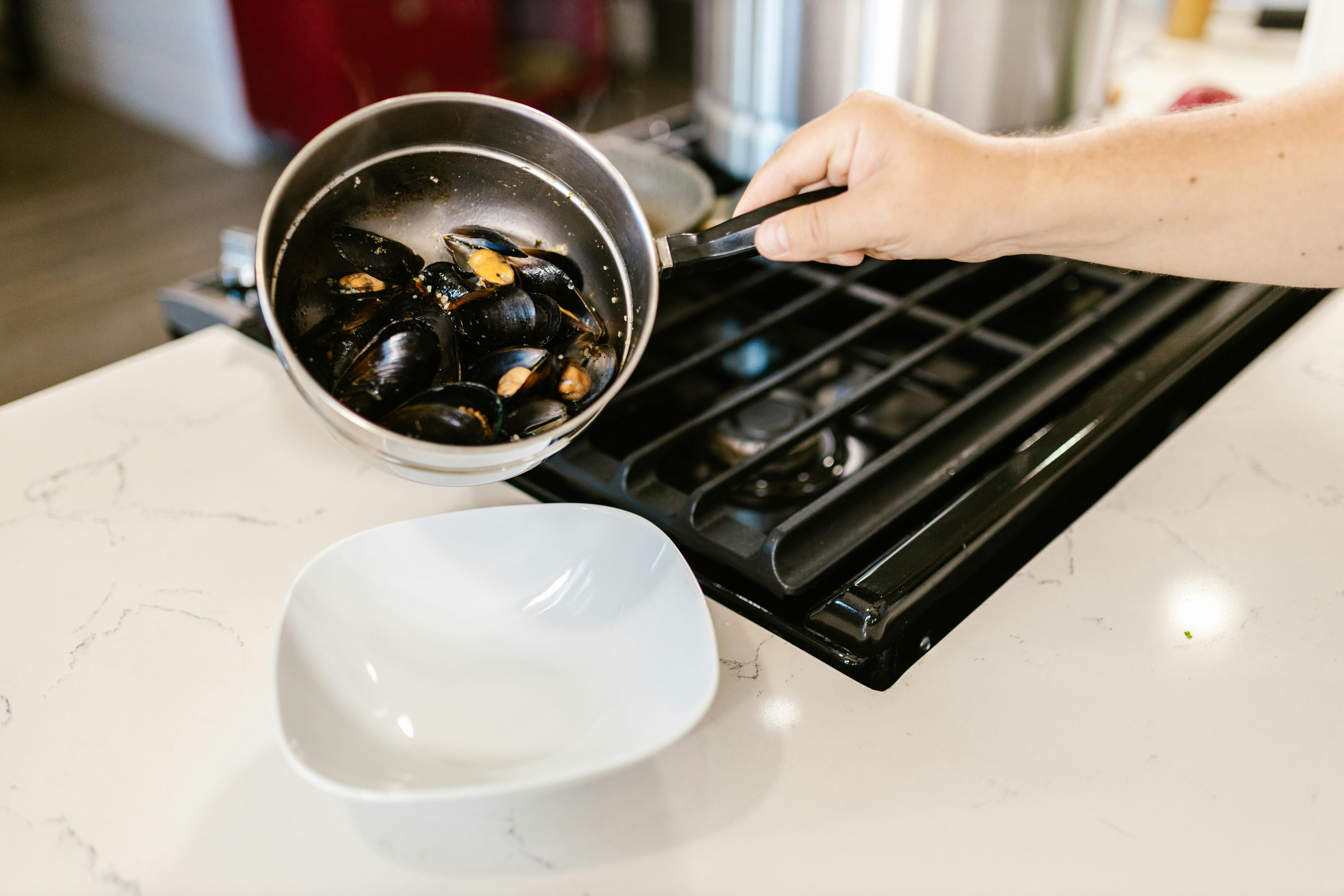 A hand preparing fresh mussels in a pot over a stove, ready to serve.
