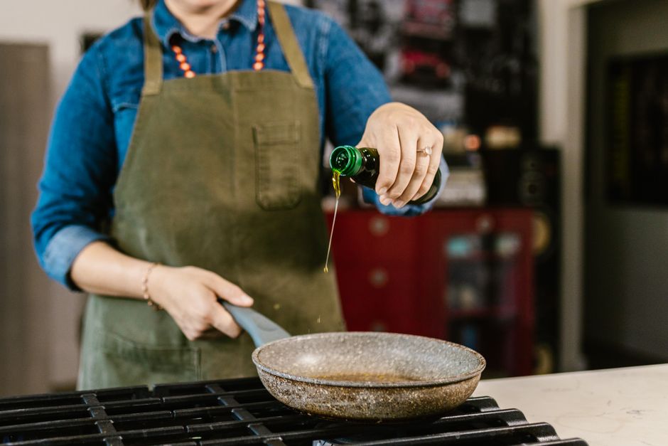 Woman in an apron pouring olive oil into a frying pan in kitchen.