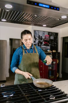 A woman chef wearing an apron pours oil into a pan in a stylish kitchen setting.