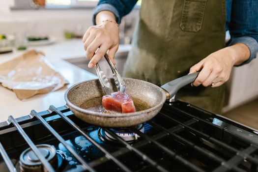 Close-up of a person cooking fresh tuna steak in a pan on a gas stove.