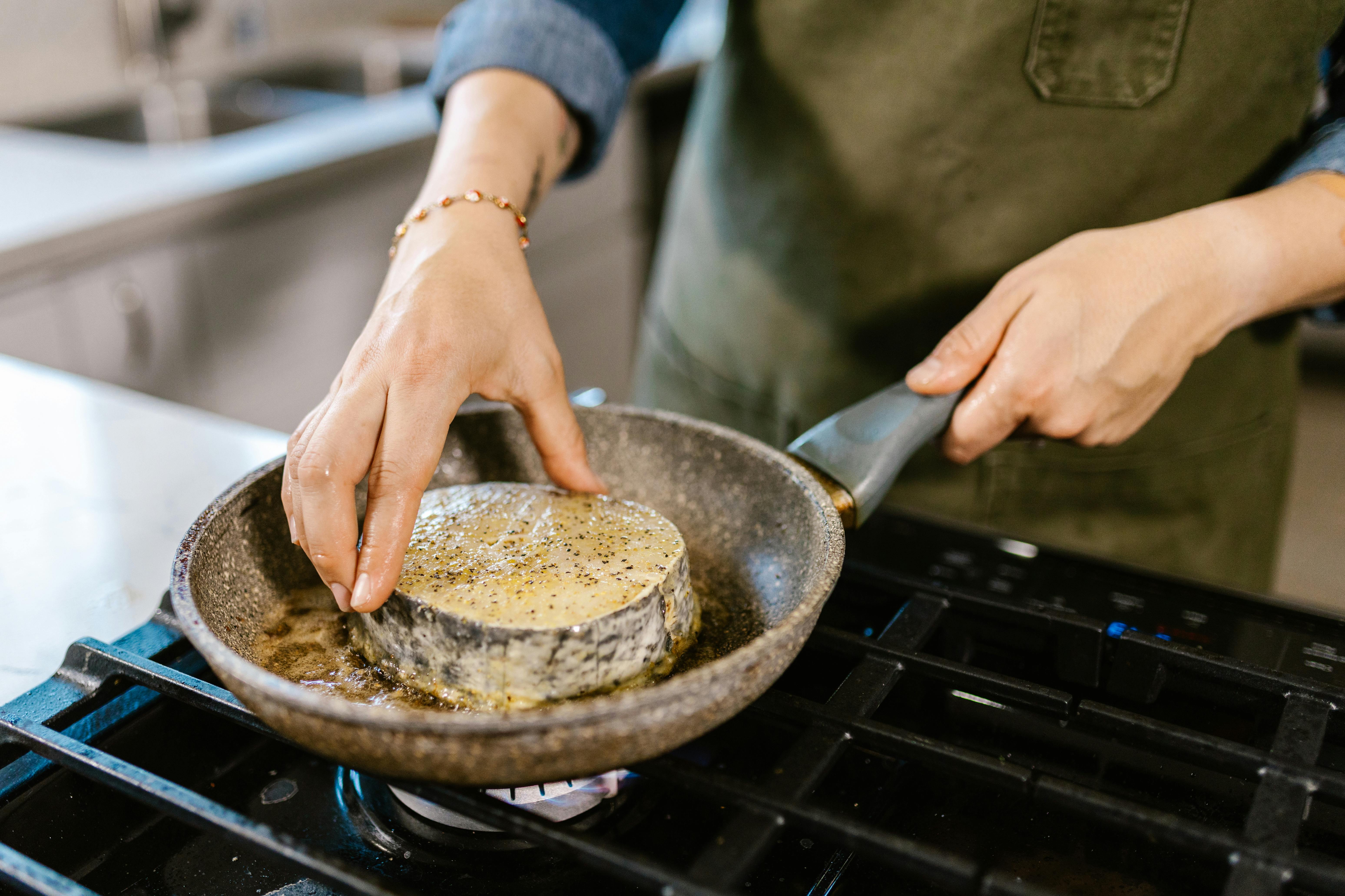 Close-up of hands cooking a seasoned fish steak in a skillet on a gas stove.