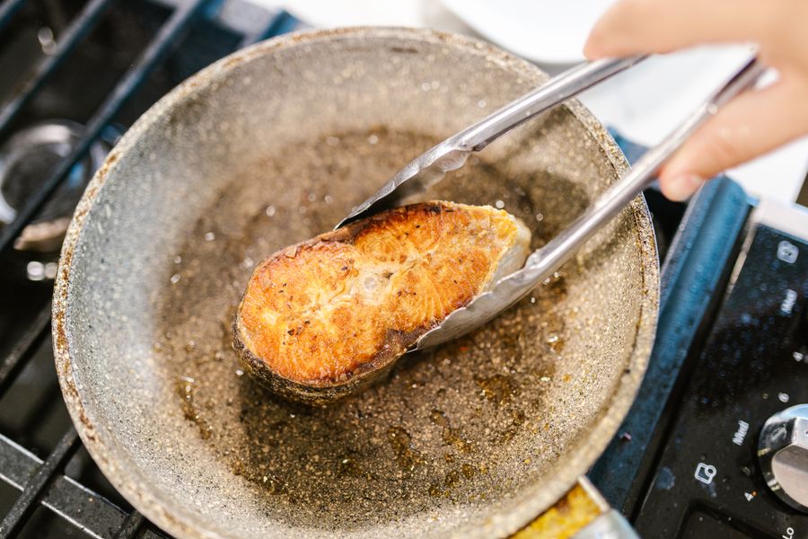 Close-up of salmon being cooked with tongs in a hot frying pan on a stove.
