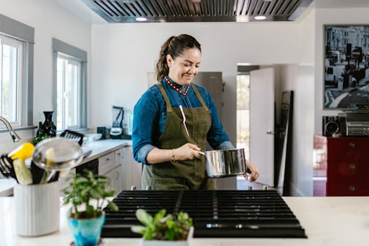 Smiling woman wearing apron cooks in a modern kitchen, holding a pot on the stove.