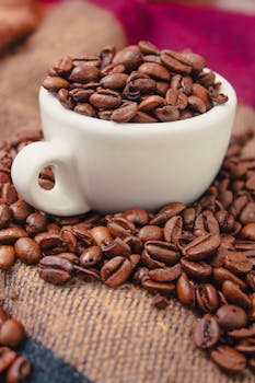 Close-up of roasted coffee beans overflowing from a white mug on a burlap sack.