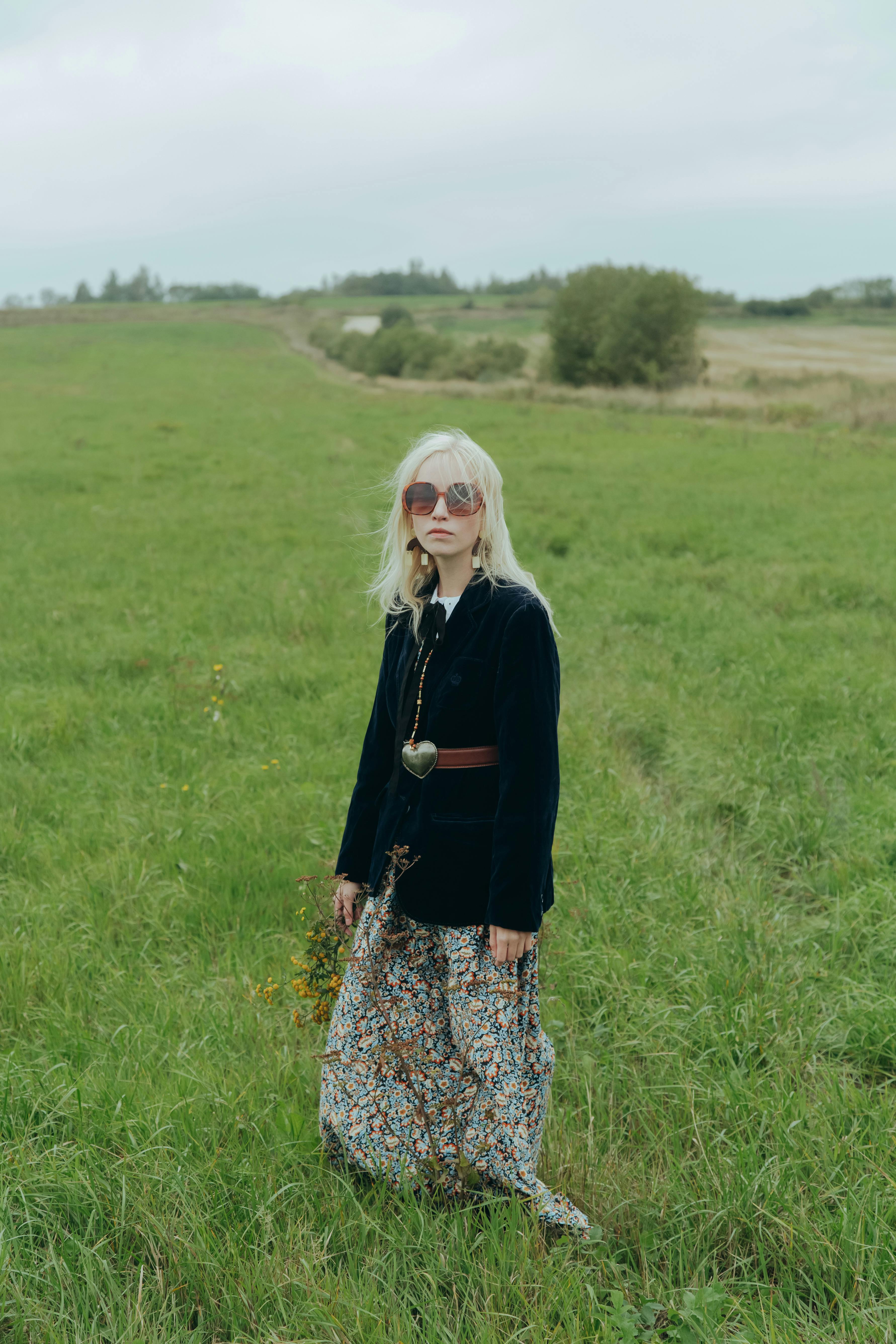 Woman in vintage attire stands in a green field with a peaceful expression.