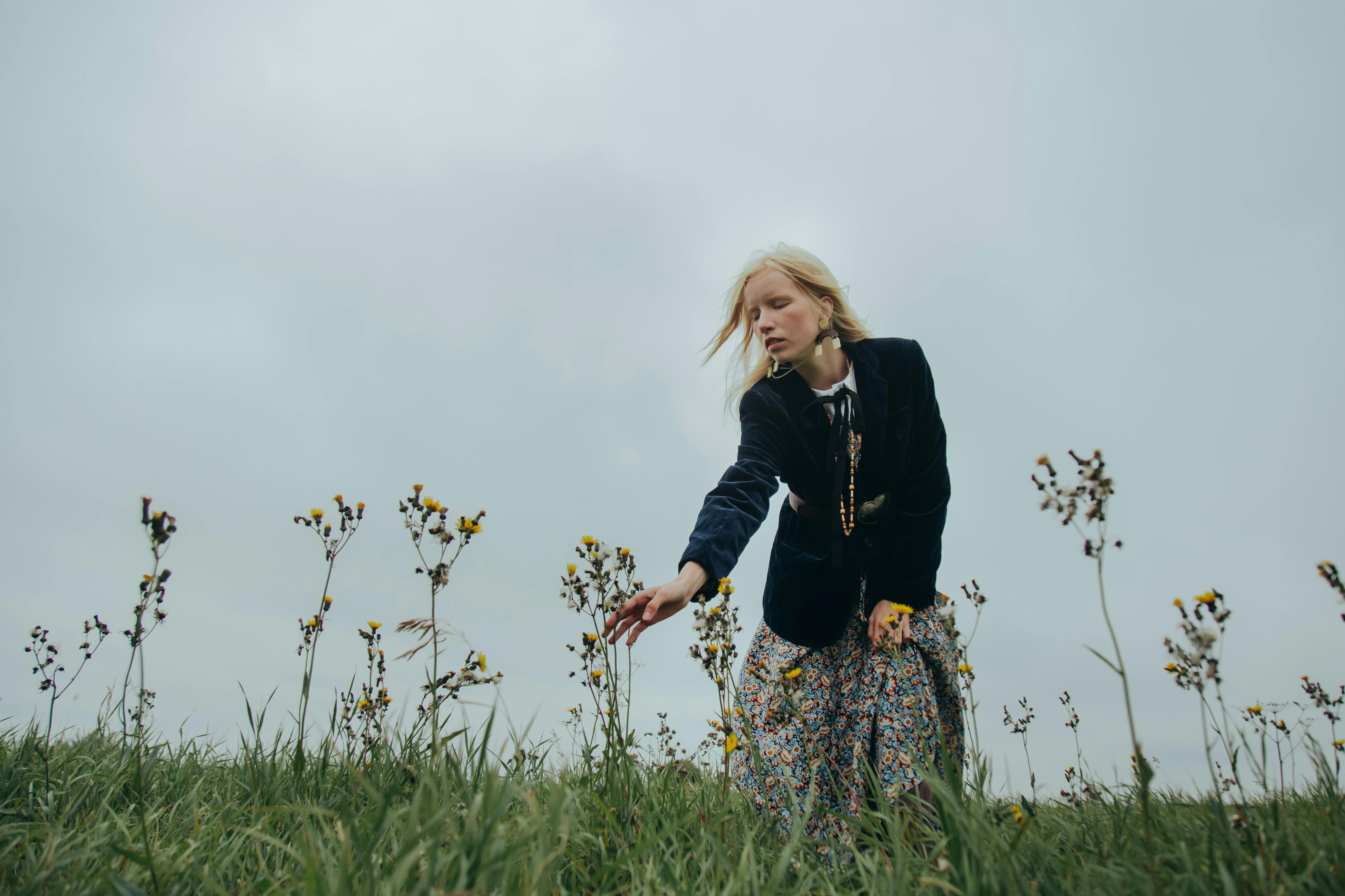 A woman in a floral dress picking wildflowers in an open field under a cloudy sky.