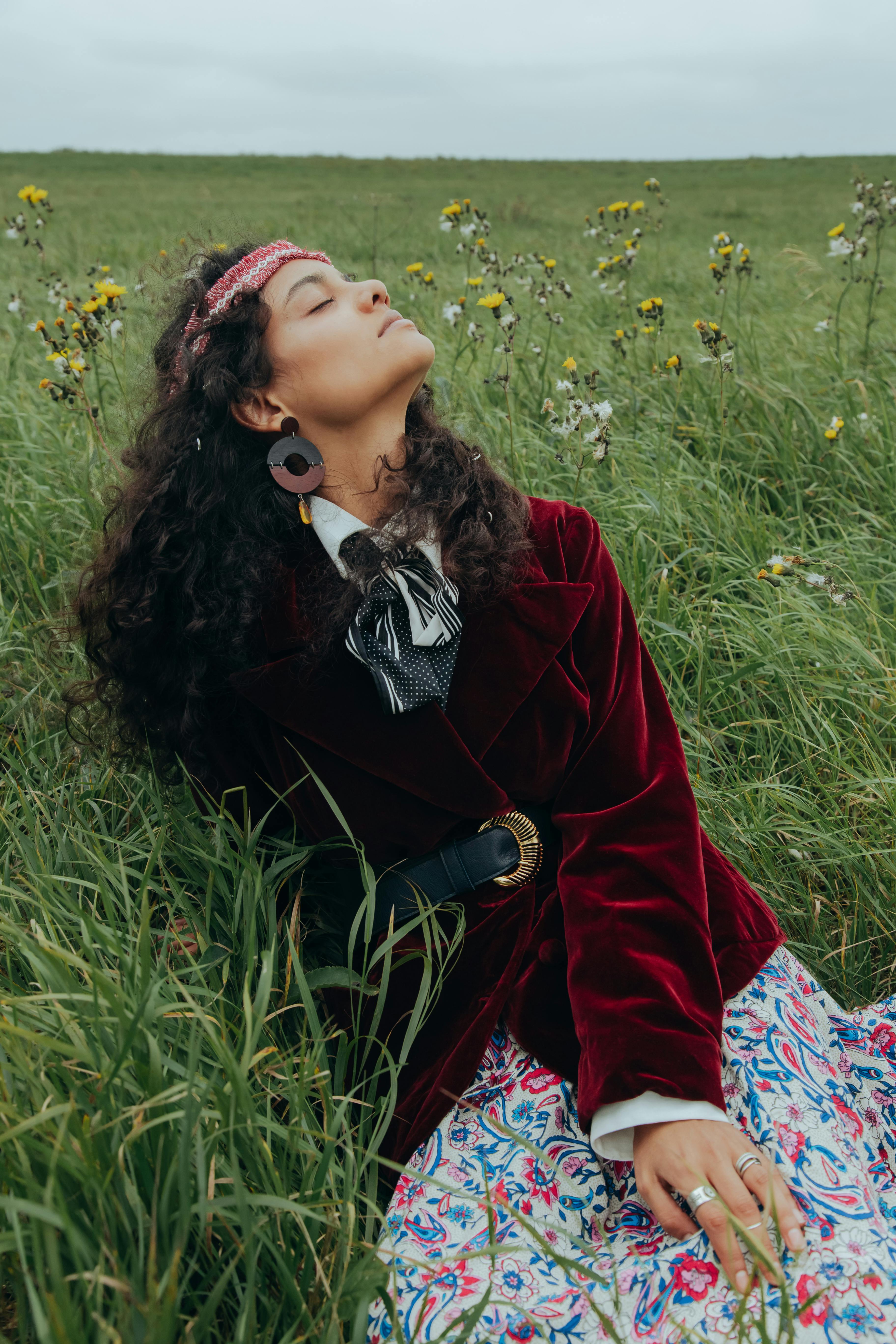A woman in a red coat sits peacefully in a grassy field. Her serene pose and closed eyes create a calming scene.