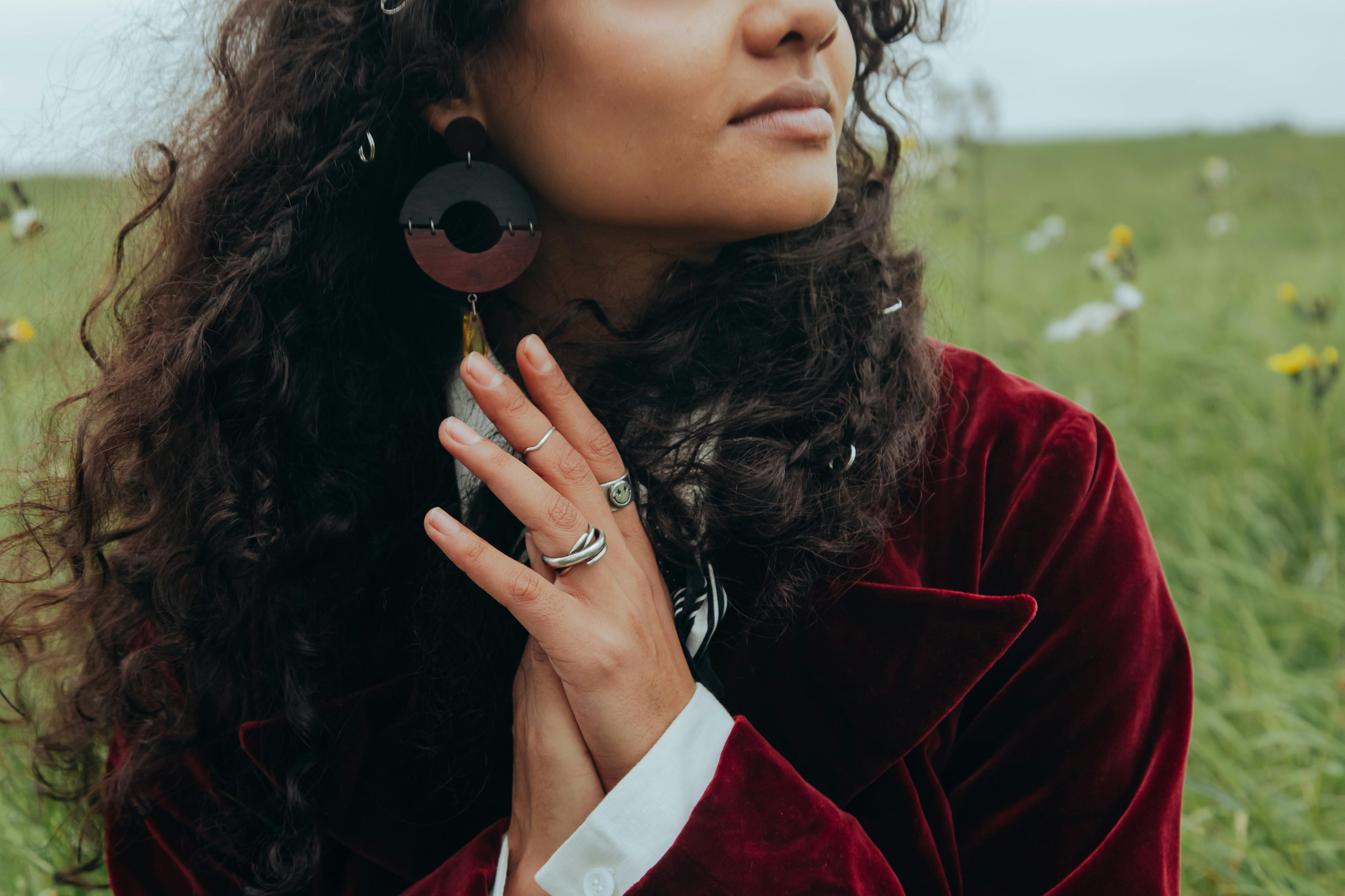 A woman with curly hair wearing vintage earrings and rings, standing in a grassy meadow.