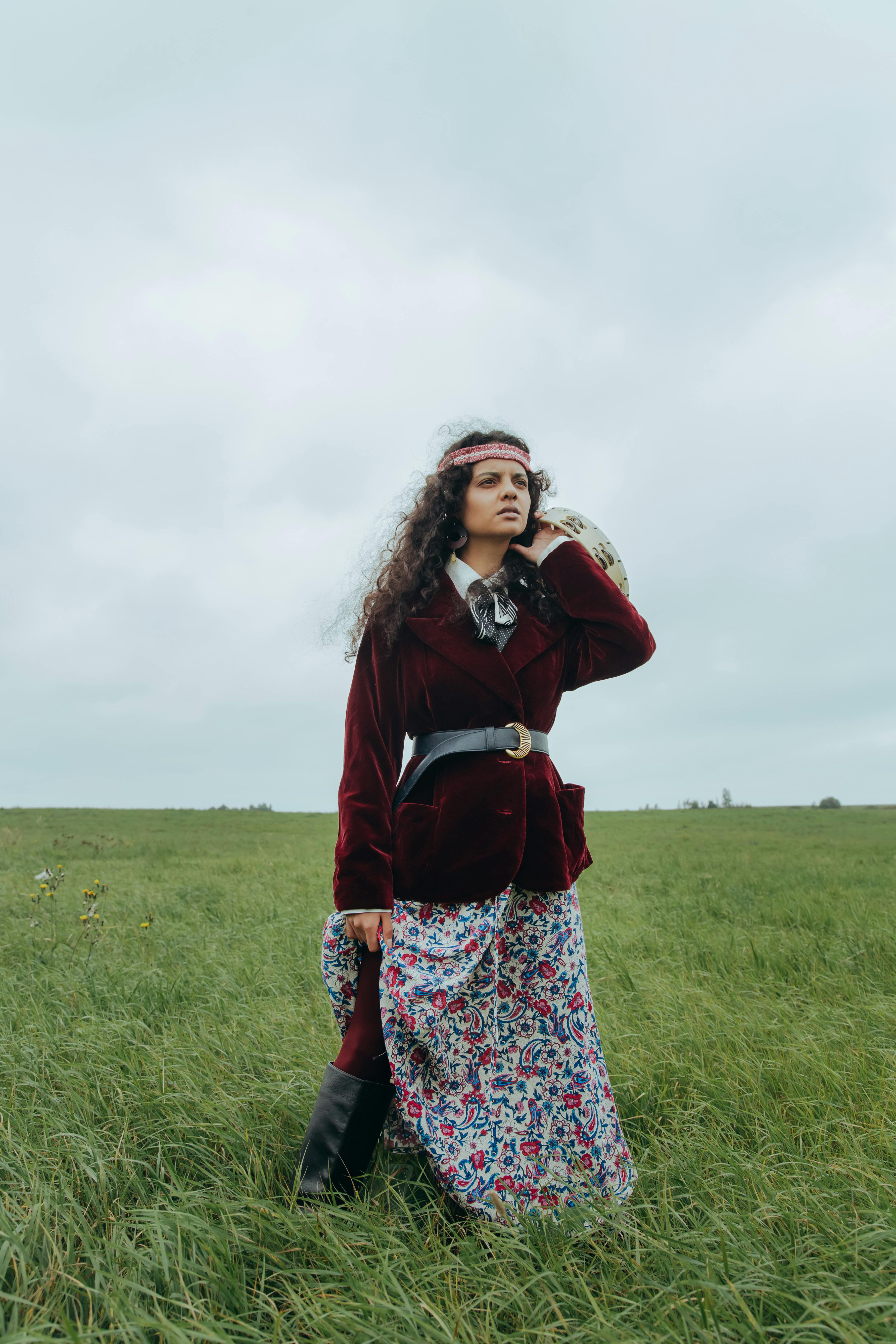 Stylish woman with a bohemian outfit stands in a green field under a cloudy sky.