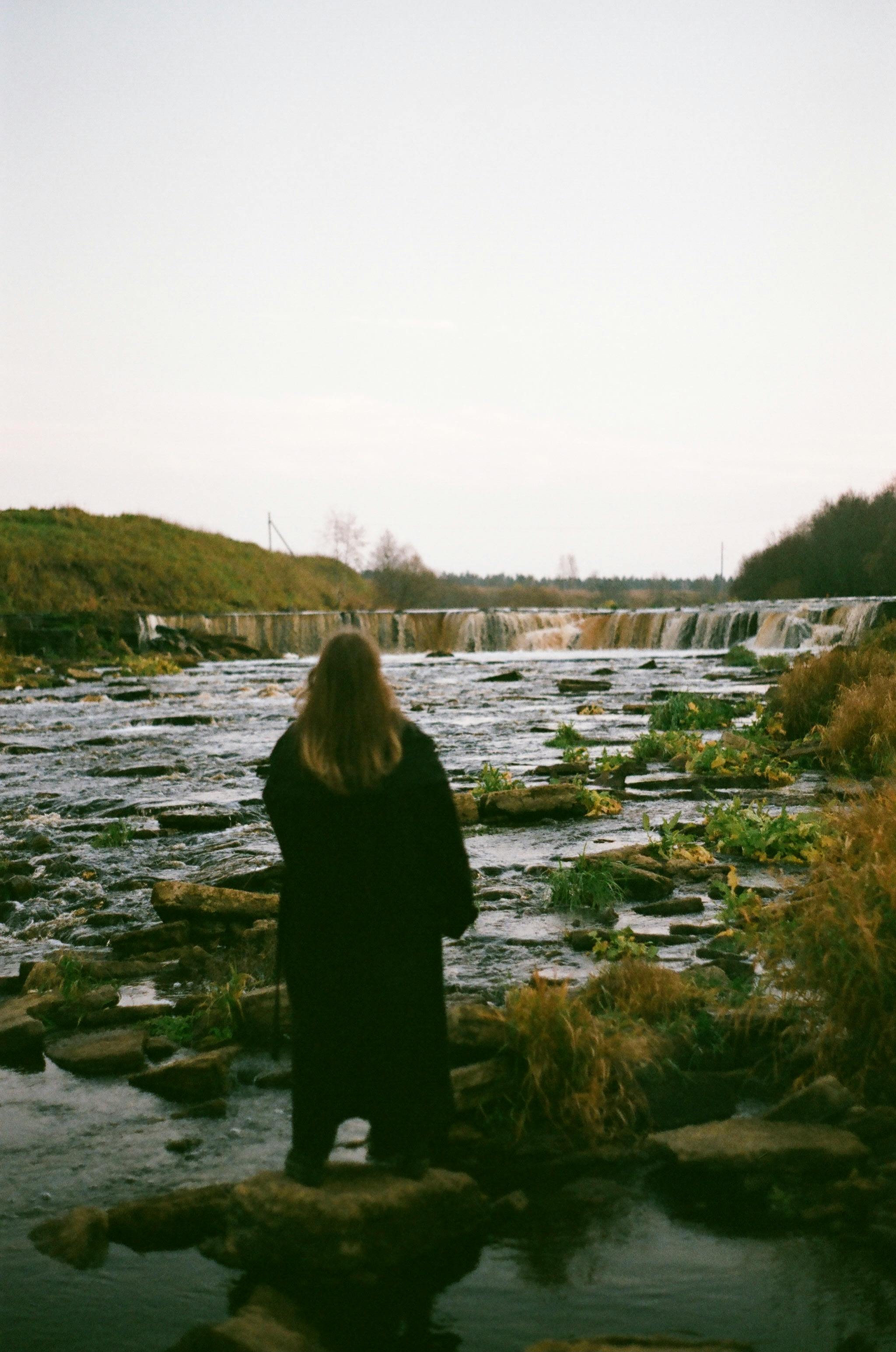 A woman stands by a tranquil waterfall, enjoying a peaceful natural landscape.