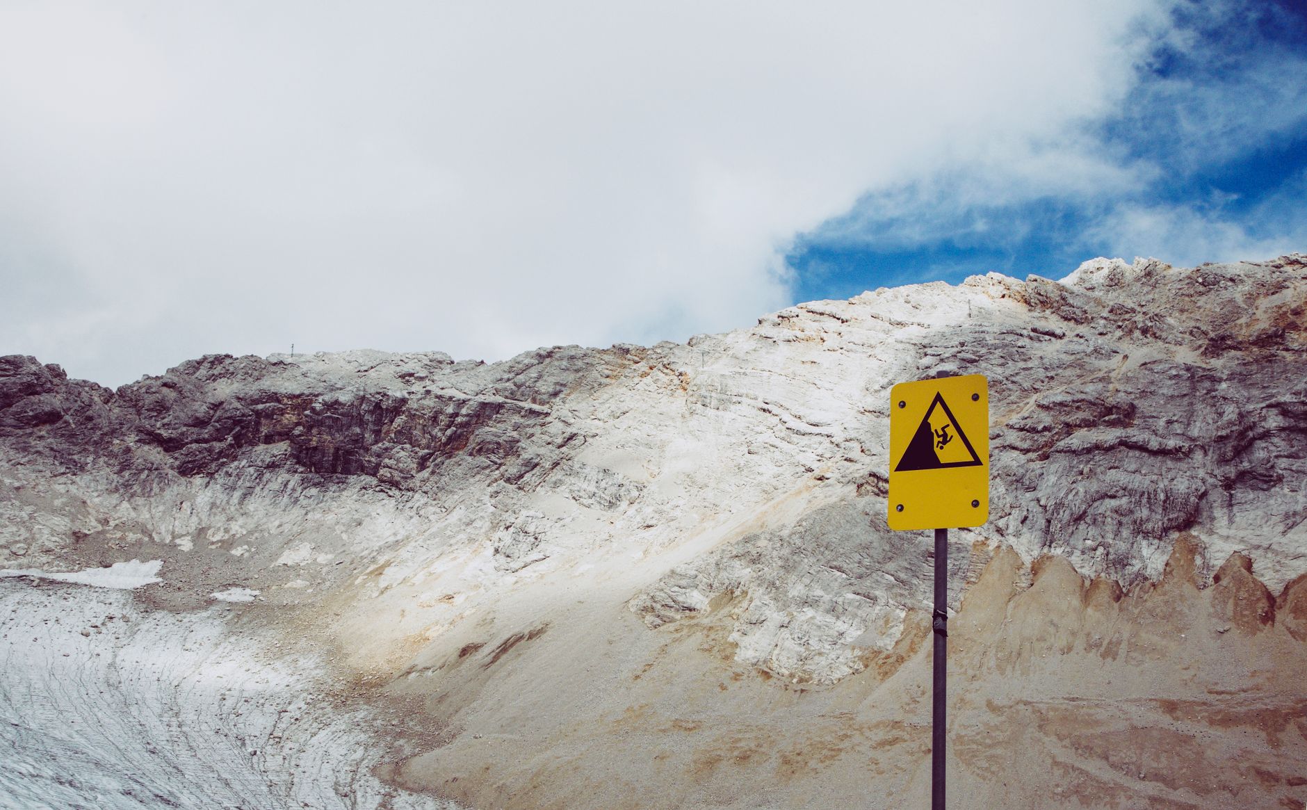 Mountainous terrain with a caution sign against falling rocks under a cloudy sky.