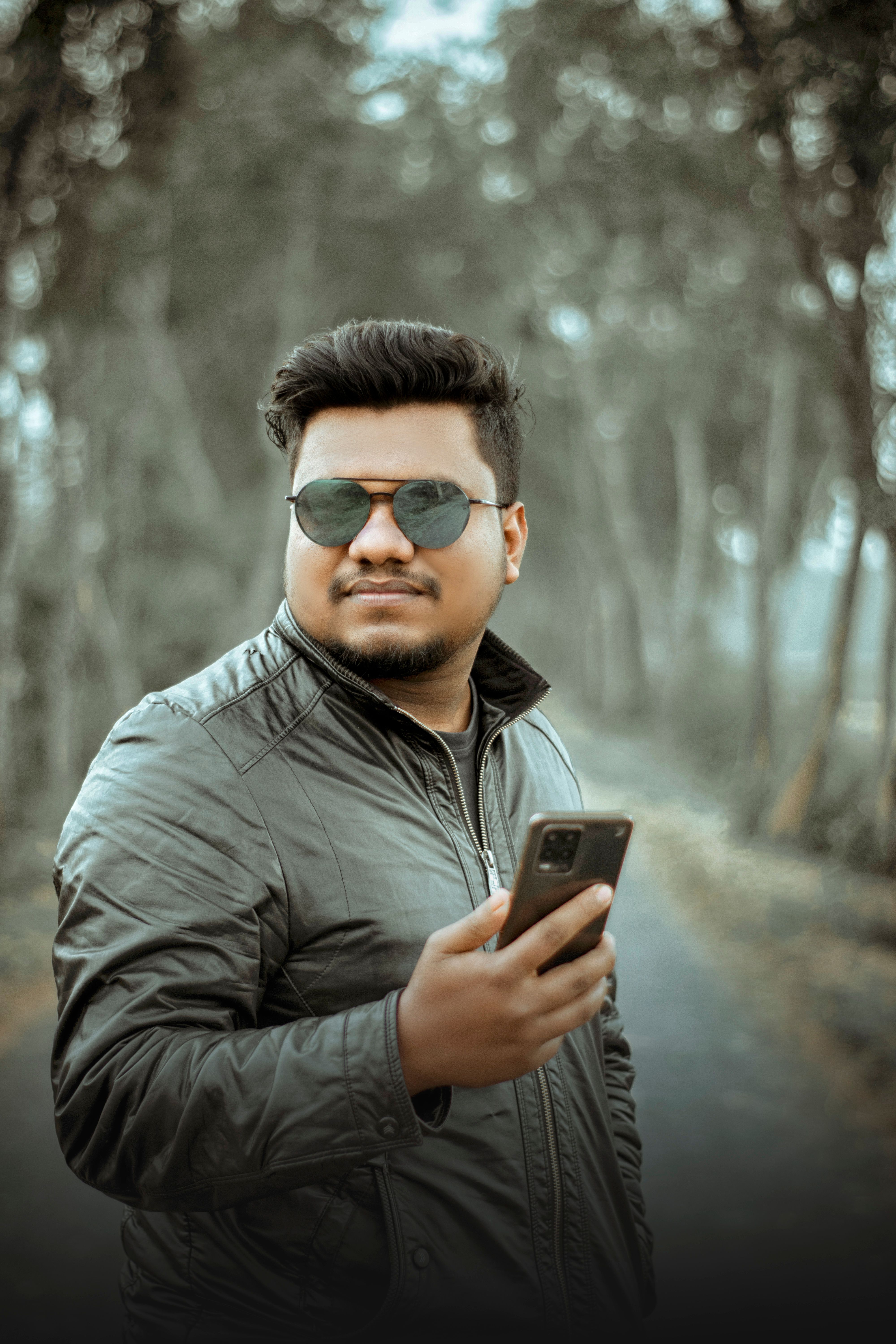 Portrait of a man in a leather jacket with sunglasses holding a smartphone on a forest path.