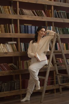 Woman in a library leaning on a ladder with books, wearing cozy attire.