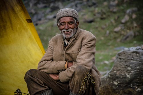 Улыбающийся человек в серой вязаной шапке, сидя возле серой скалы