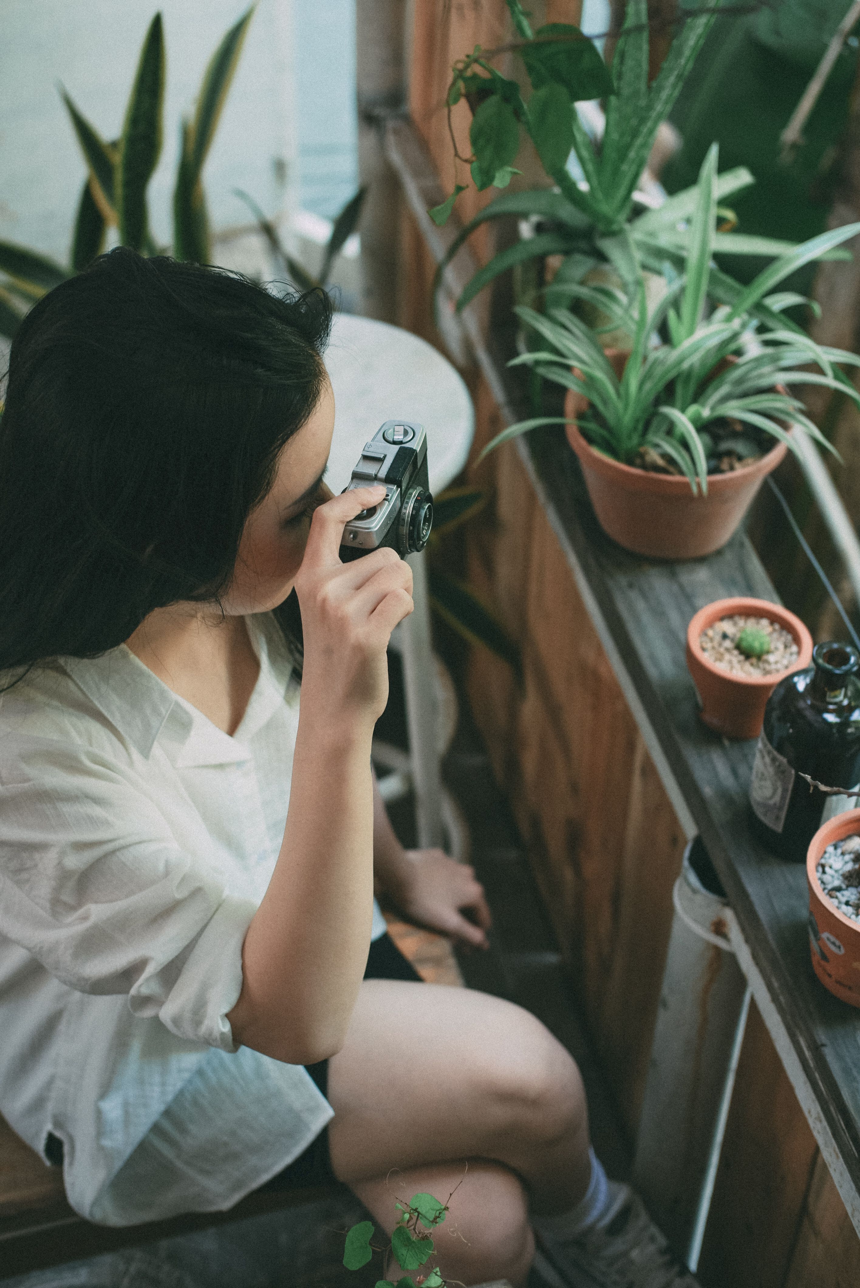 A woman using a digital camera to photograph houseplants on a wooden shelf.