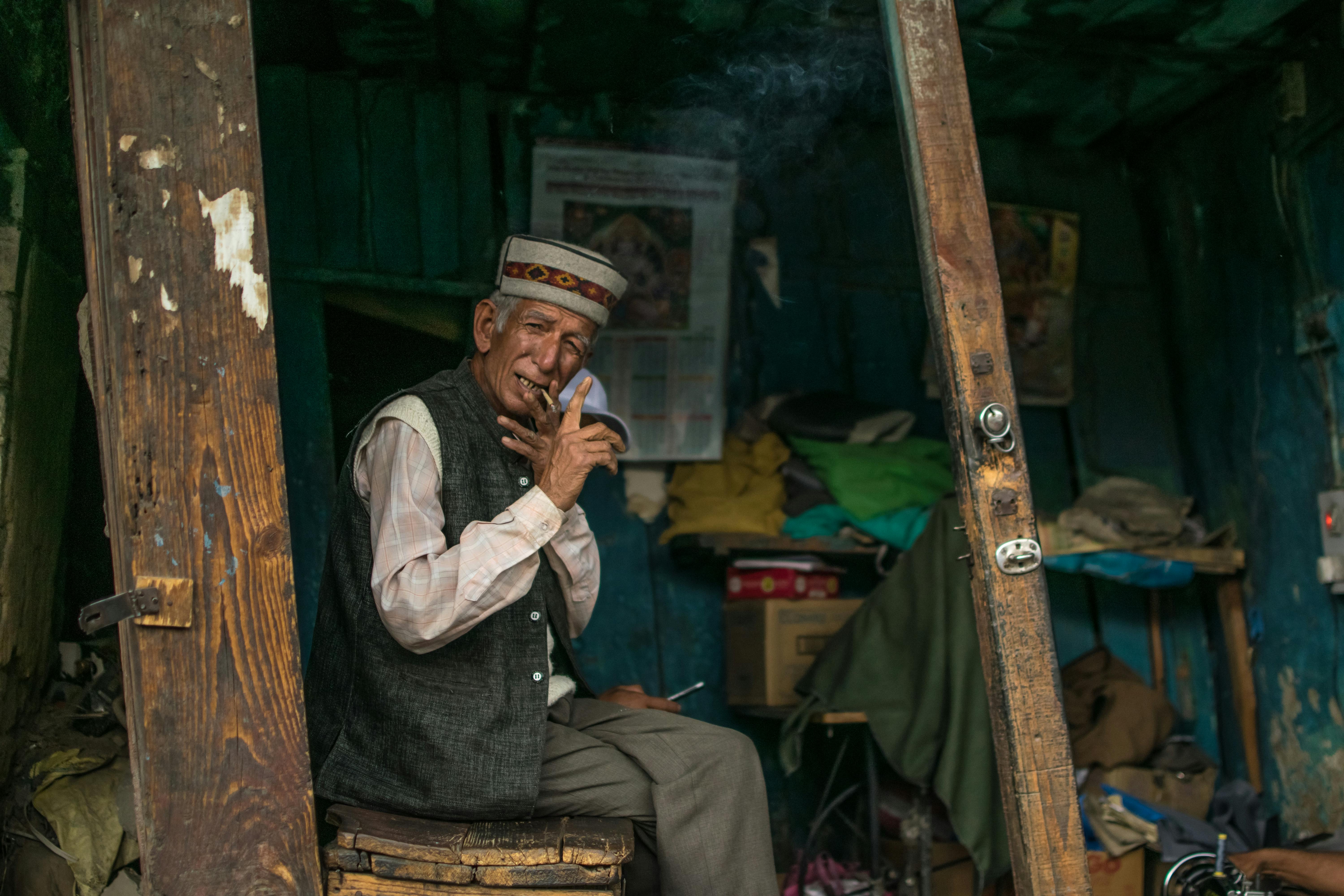An elderly man in traditional attire smokes in a rustic wooden hut, exuding cultural charm.