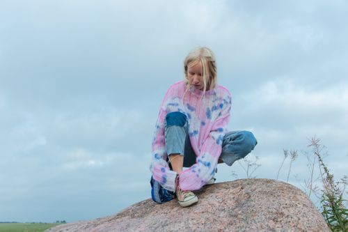 Free Blonde woman in tie-dye sweater sitting on a rock in a serene outdoor setting. Stock Photo