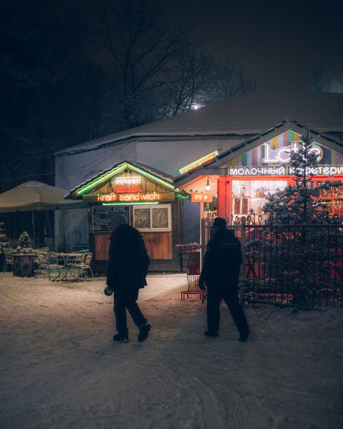 Free Winter night view of illuminated streetside cafe with people walking on snow. Warm and inviting ambiance. Stock Photo